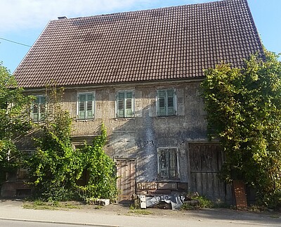 Frontansicht Das Bild zeigt die Frontansicht eines alten, offensichtlich unbewohnten Hauses. Die Fensterläden sind geschlossen, vor dem Haus wuchern große Gebüsche.