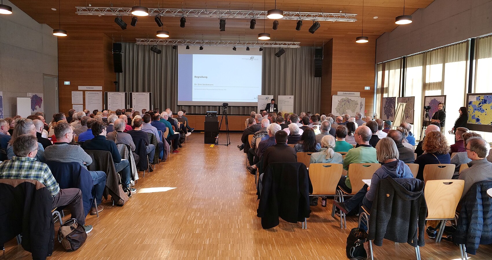 Man blickt von hinten in einen voll besetzten Veranstaltungsraum. Ganz vorne ist das Plenum mit einem Sprecher zu erkennen.